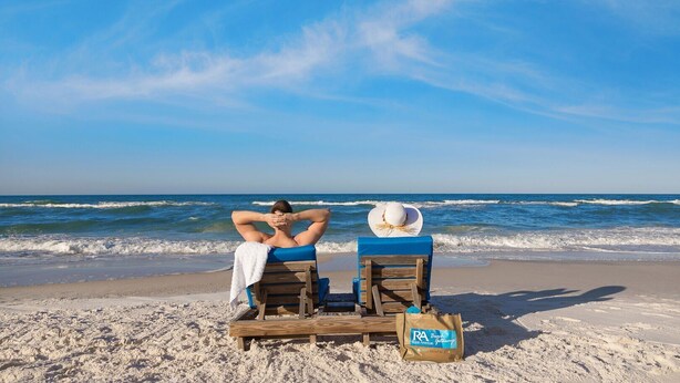 A beach scene with two wooden lounge chairs, one with a towel and a bag, and the ocean in the background. - 3BR 3.5BA @ EMERALD BEACH! SLEEPS 10! (Panama City Beach)