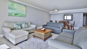 A living room with a light-colored sofa, a blue sofa, a wooden coffee table, and a dining area with wooden chairs and a table. - 3BR 3.5BA @ EMERALD BEACH! SLEEPS 10! (Panama City Beach)