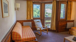 Ein gemütliches Zimmer mit einer hölzernen Bank, einem Schaukelstuhl und einem kleinen Tisch. Große Fenster bieten Blick auf eine gebirgige Landschaft.