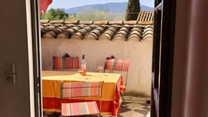 An open door to a patio with a checkered floor, a table with a yellow tablecloth, striped chairs, and a red umbrella. - Maison "molière" (Ferrals-les-Corbières)