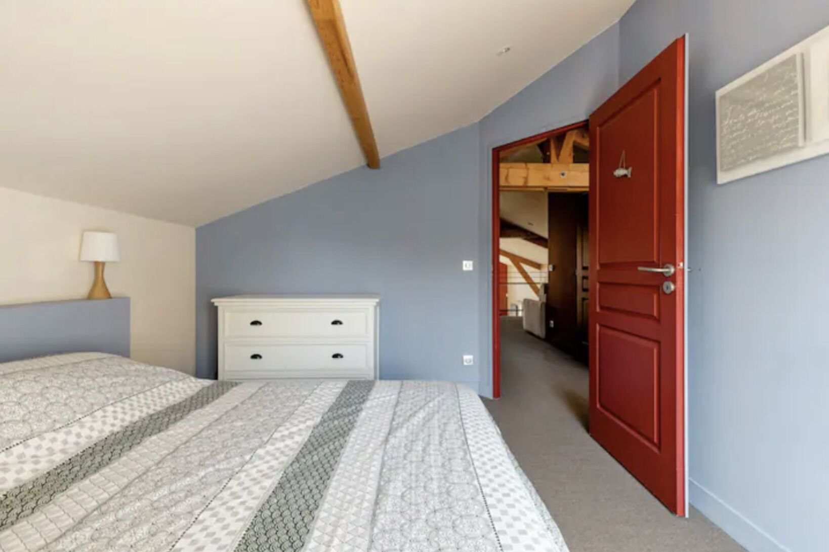 A bedroom with a bed, a dresser, a red door, and a hallway leading to another room.