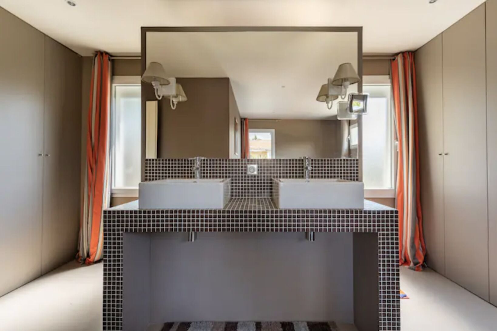 A modern bathroom with a large mirror, two sinks, and a window with curtains.