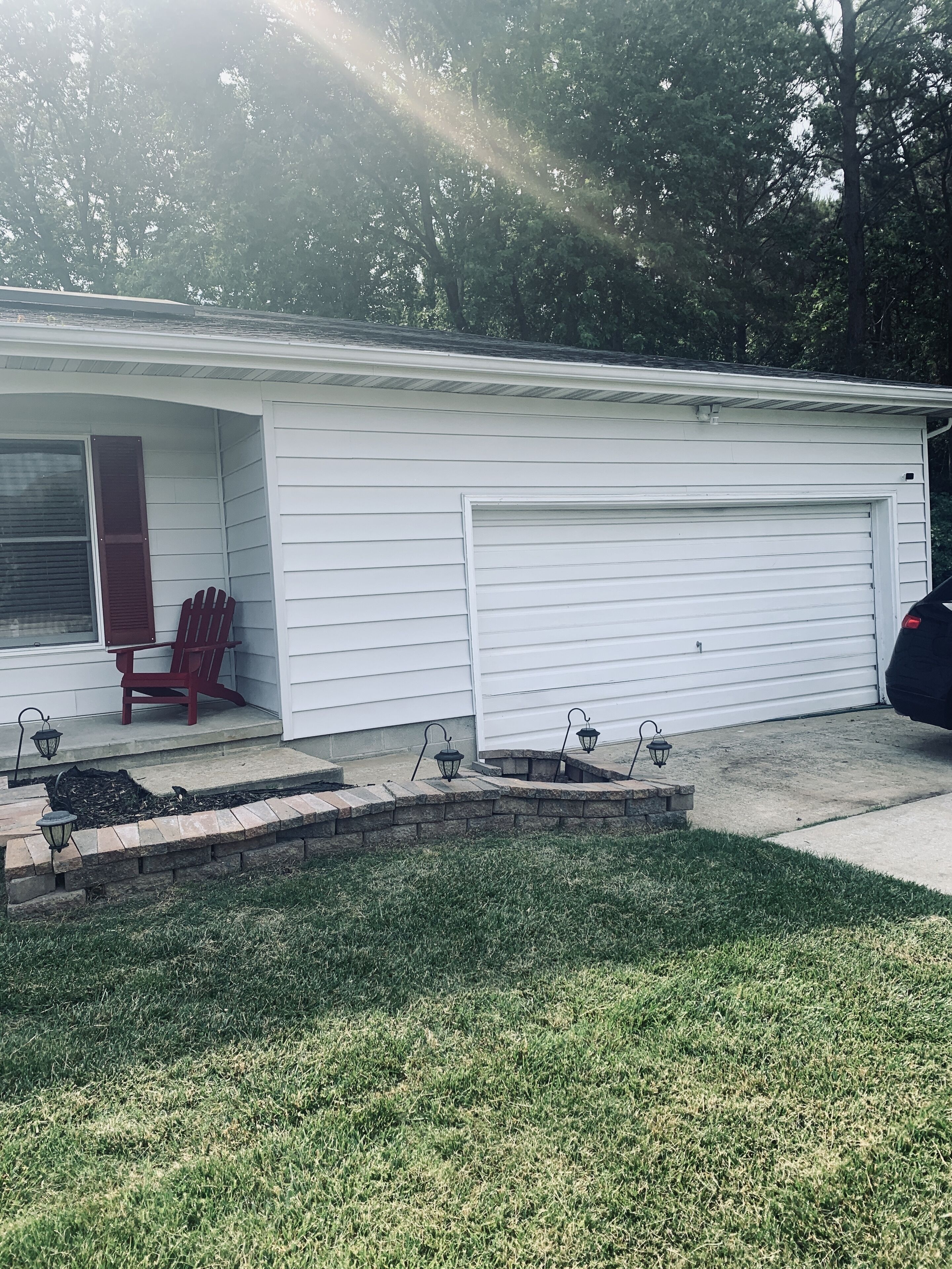 Ein eingeschossiges Haus mit Garage, einer überdachten Veranda mit einem roten Adirondack-Sessel und einem kleinen Ziegelpflanztopf mit drei schwarzen Laternen.