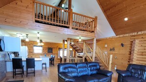 A spacious log cabin interior with a dining area, two black leather sofas, a kitchen, and a staircase to an upper level. - Log Cabin located minutes from Missouri River, airport, I-90 and downtown. (Chamberlain)
