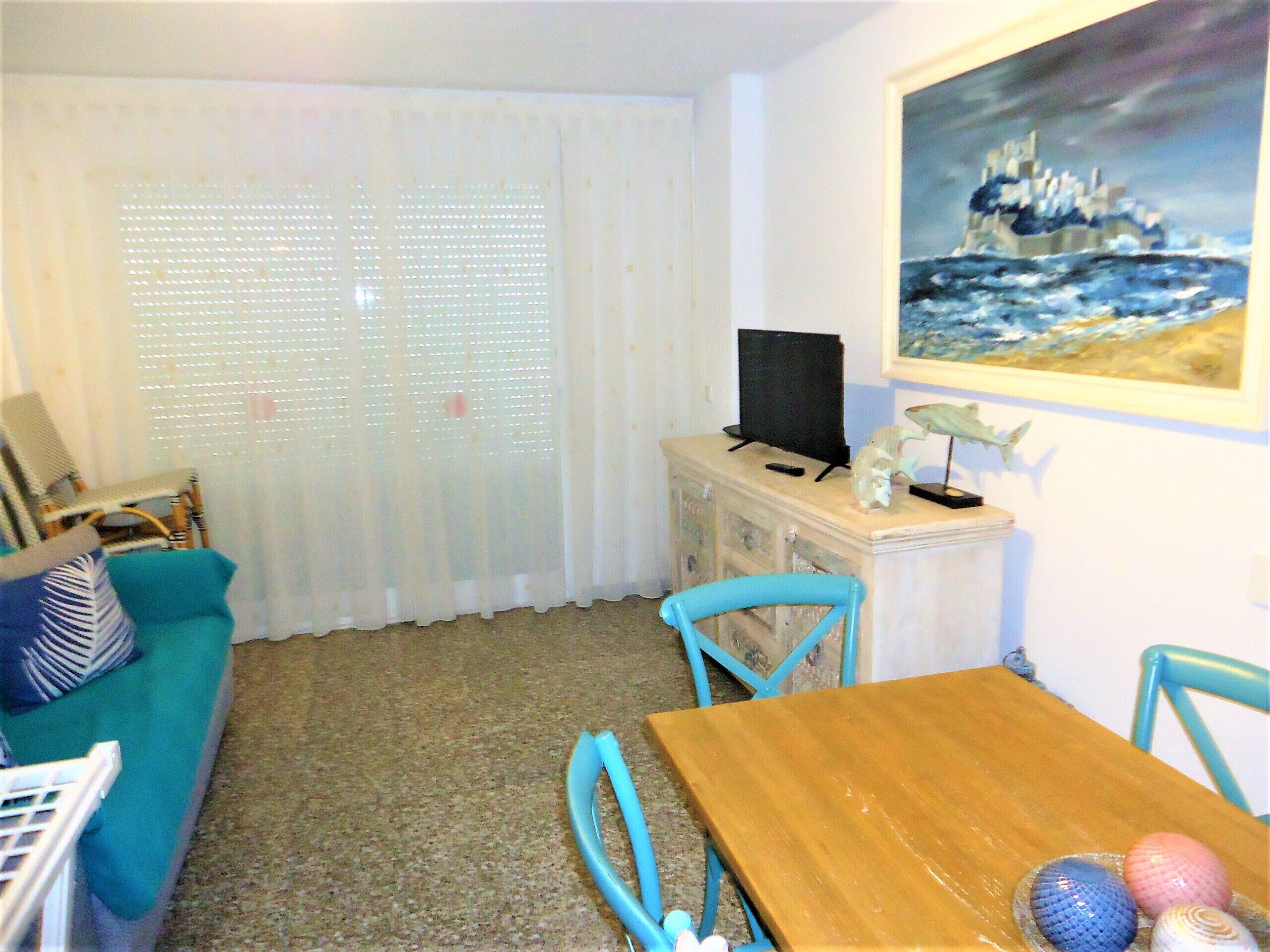Un soggiorno con un tavolo in legno, sedie blu, un divano, una TV su un mobile e un quadro appeso al muro.