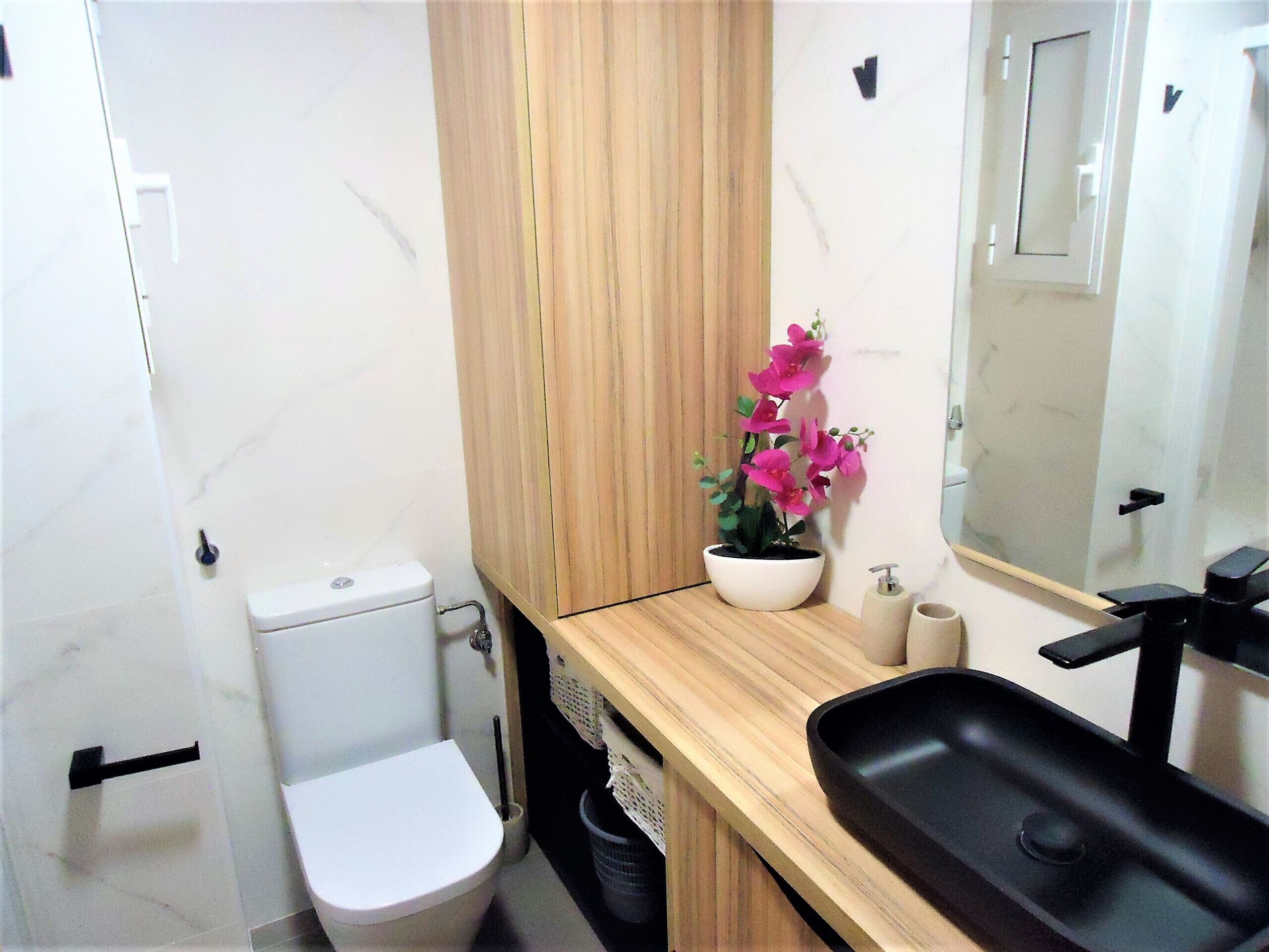 A compact bathroom with a white toilet, a wooden vanity, a black sink, and a mirror.