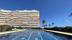 Piscina com água azul cristalina, rodeada por um edifício com várias varandas e toldos laranja.