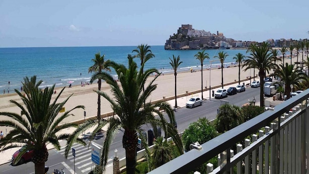 Vista costeira com praia, palmeiras e um edifício histórico do outro lado da água.