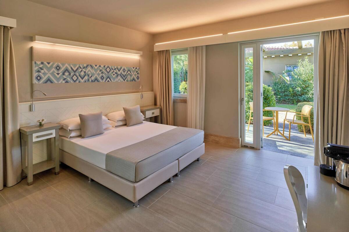Een moderne hotelkamer met een groot bed, nachtkastje, en een raam met gordijnen en uitzicht op de tuin.