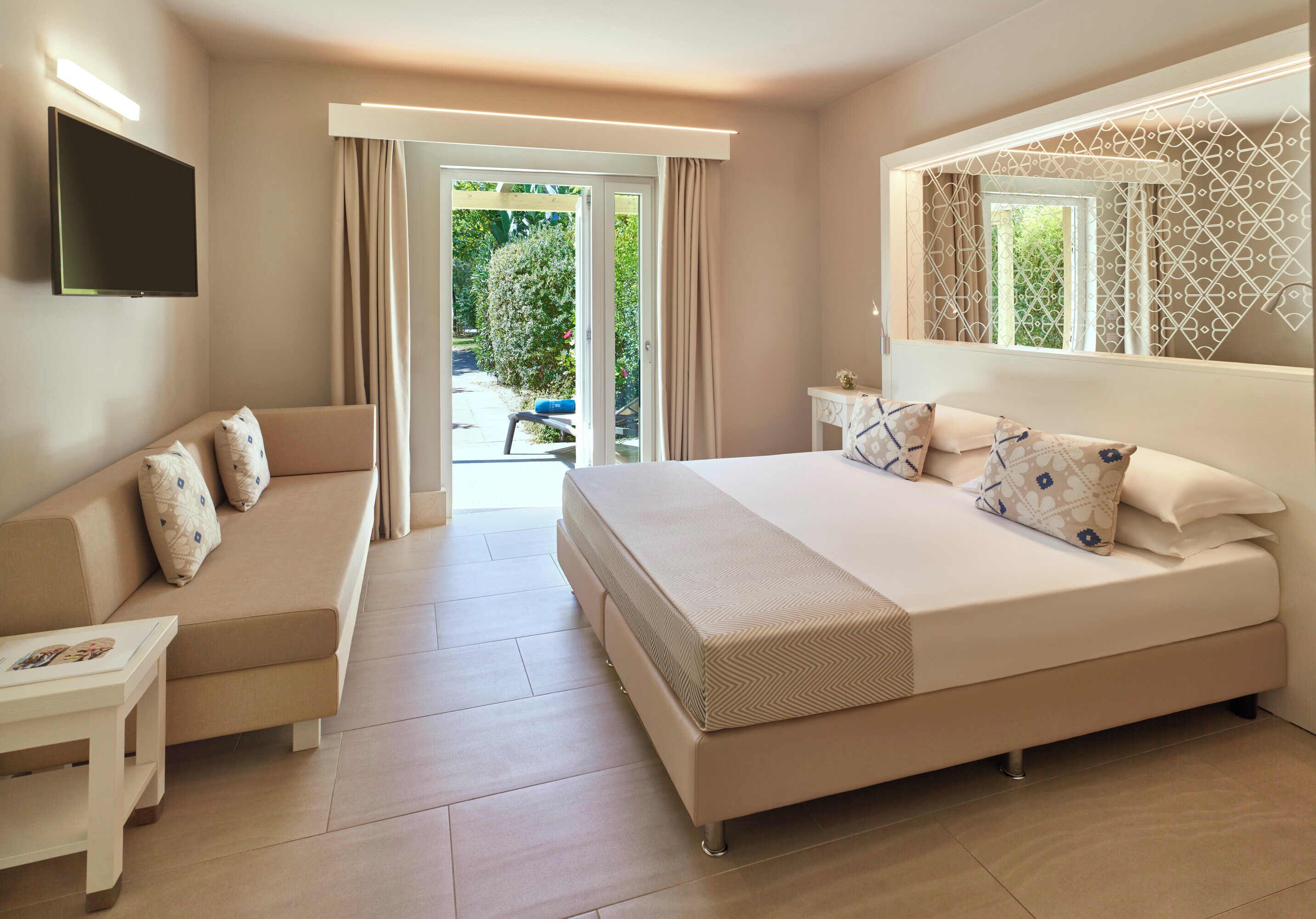 A modern hotel room with a large bed, a sofa, a TV, and a balcony with a view of greenery.