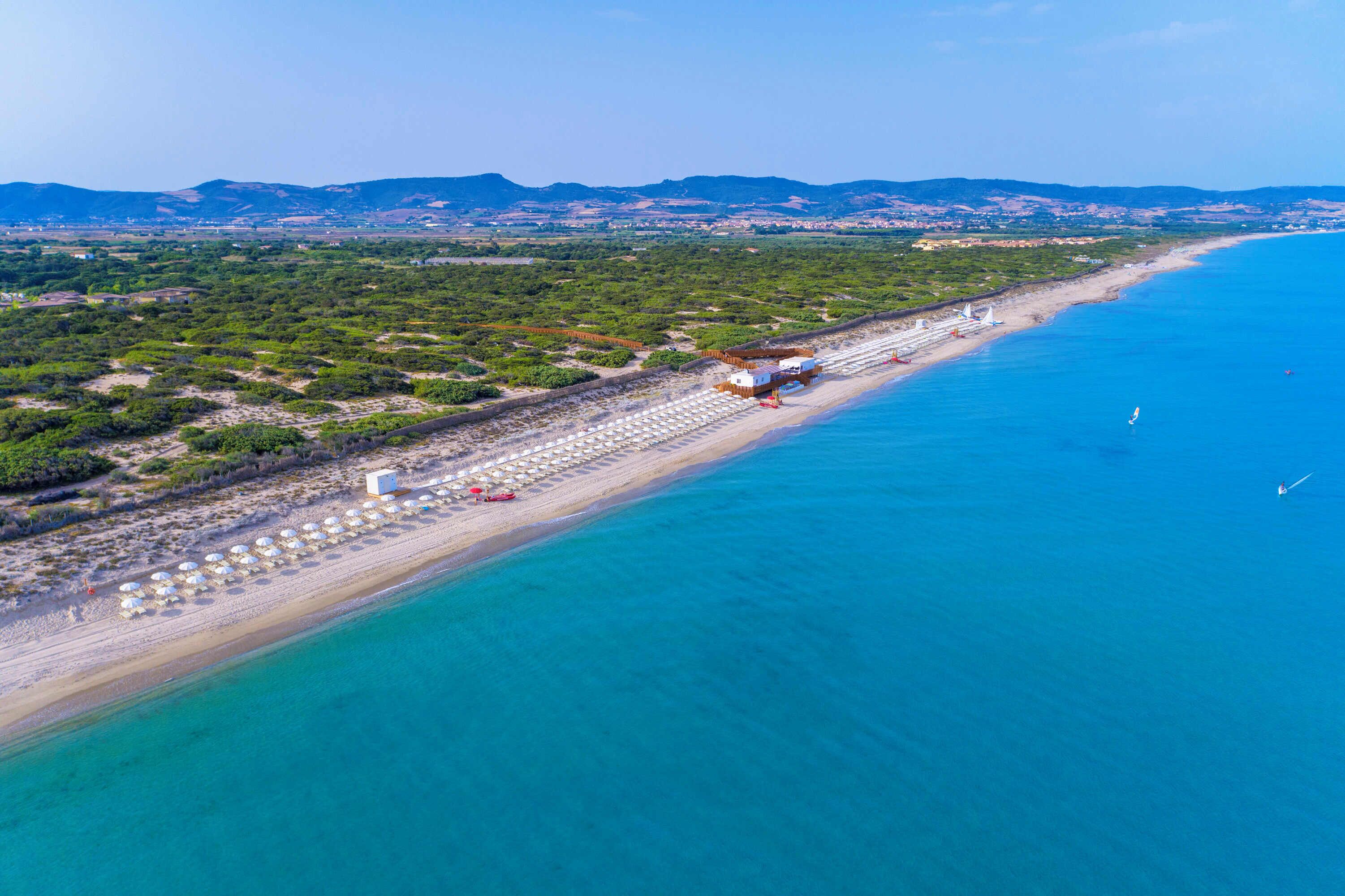 An aerial view of a beach with a row of umbrellas and beach chairs, a small building, and a clear blue sea.