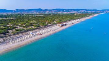 An aerial view of a beach with a row of umbrellas and beach chairs, a small building, and a clear blue sea.