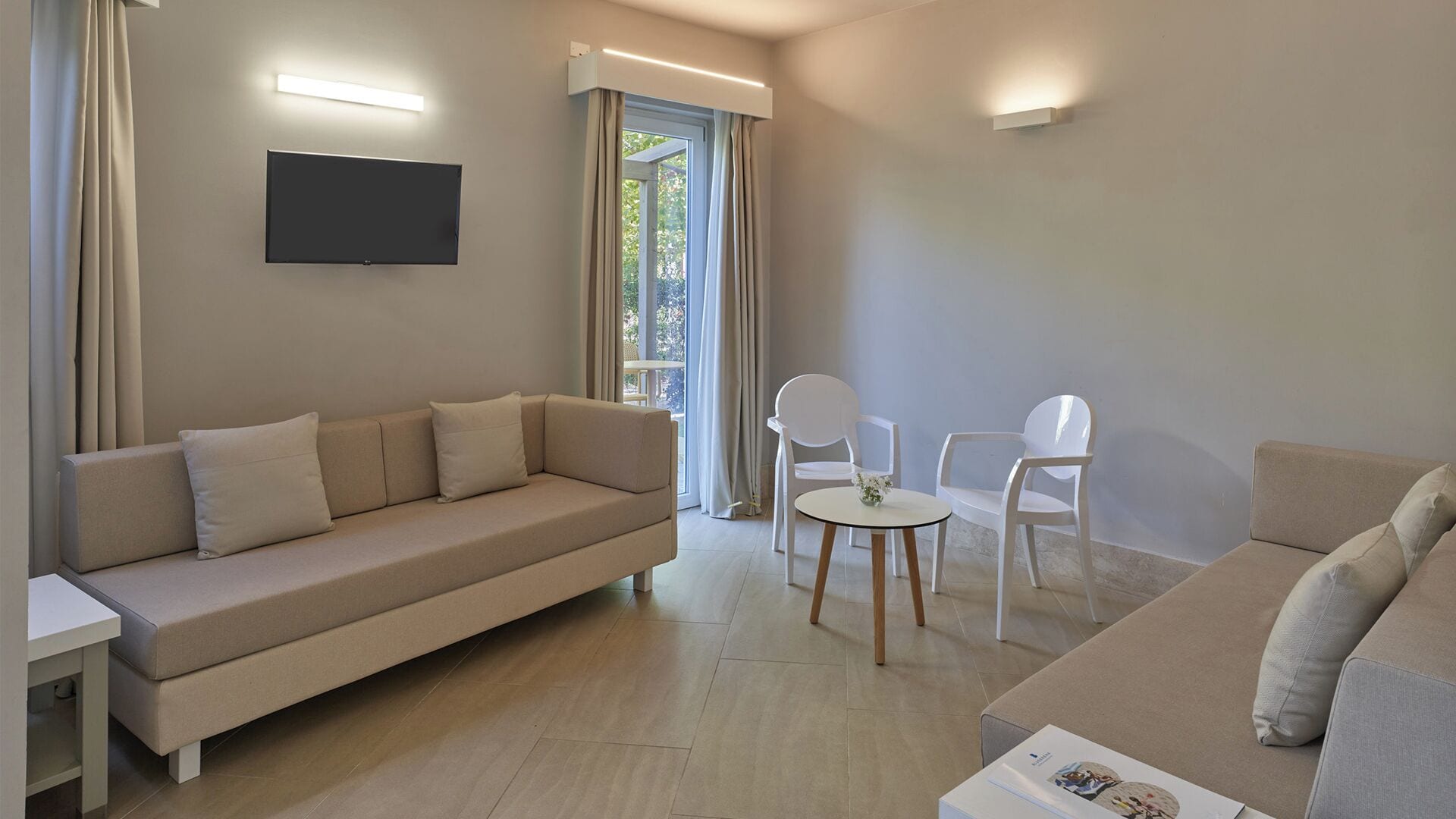 Un soggiorno moderno con un divano beige, due poltrone bianche, un tavolino rotondo e una TV a schermo piatto montata a parete.