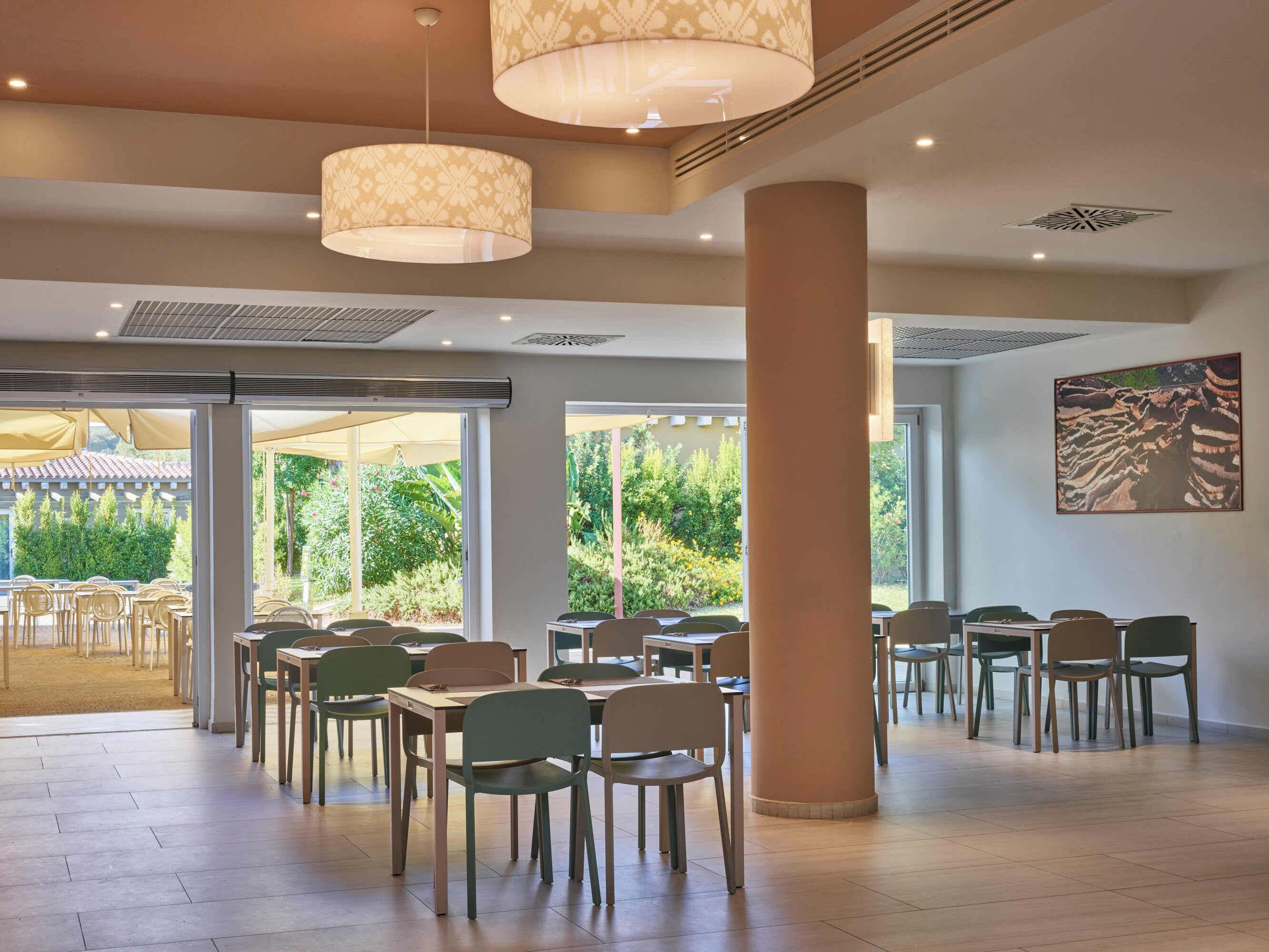 Un comedor amplio con varias mesas y sillas, grandes ventanales que dan a zonas verdes y un techo con lámparas decorativas.