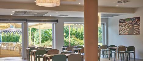 A spacious indoor dining area with multiple tables and chairs, large windows offering a view of greenery, and a ceiling with patterned lamps.