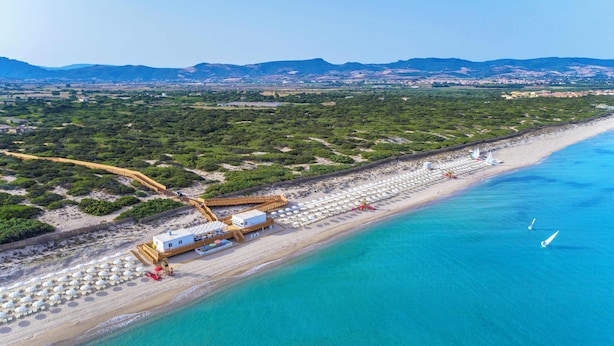 Vue aérienne d’un complexe hôtelier en bord de mer, avec une longue plage, une promenade en bois et un bâtiment à la façade blanche.
