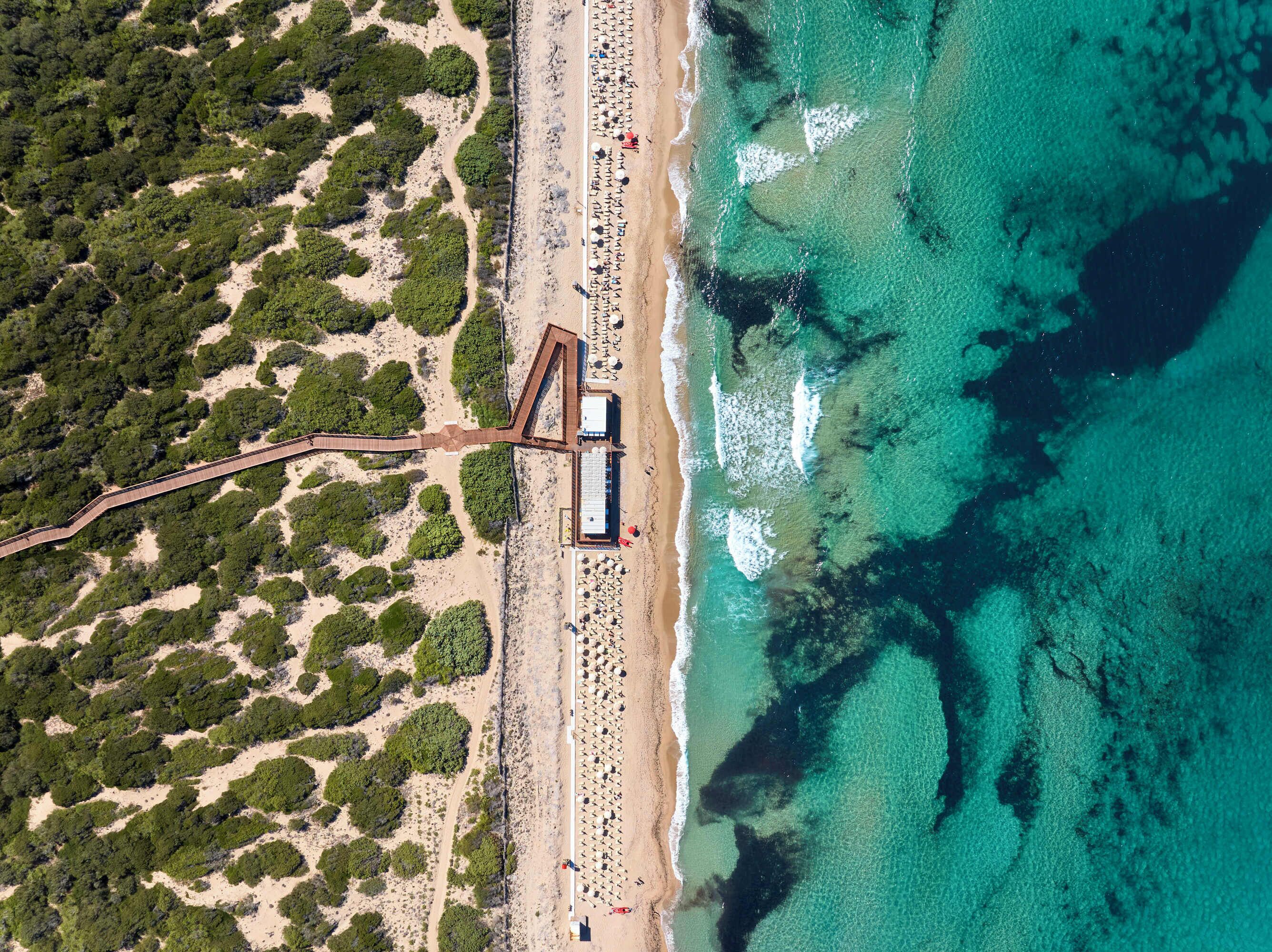 Vista aerea di una spiaggia con una passerella in legno, ombrelloni e un edificio a tetto piatto.