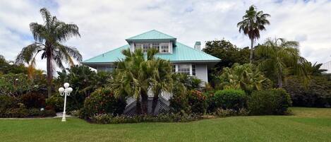 A two-story house with a green roof, surrounded by lush greenery and palm trees.