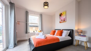 Un dormitorio moderno con una cama negra, ropa de cama naranja, una mesita de noche blanca con libros, una ventana con cortinas y un cuadro en la pared.