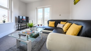Una sala de estar moderna con una mesa de centro de vidrio, un sofá de cuero negro con cojines amarillos, un televisor de pantalla plana y un ventanal con vistas.