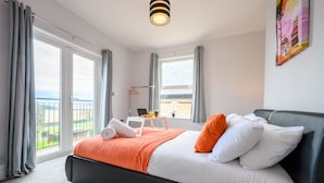 Una habitación de hotel moderna con una cama grande, un balcón con vistas y una pequeña mesa con una computadora portátil y una taza.