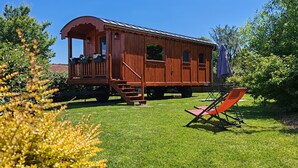 En trævogn med veranda og vinduer, placeret på en græsplæne med en stol og et lilla håndklæde i nærheden.