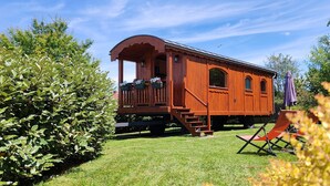 En trætogsvogn med veranda og vinduer, placeret på en græsplæne med en stol og planter i nærheden.
