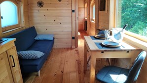 Indretning i en hyggelig hytte med trægulv, en blå sofa, et spisebord med stole og et vindue med udsigt til grønne omgivelser.