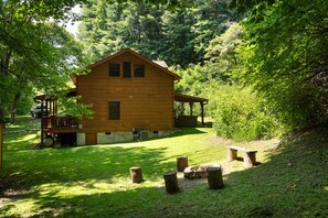 A wooden cabin with a porch and a covered area, surrounded by lush greenery.