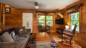 Accogliente interno di una baita con arredamento in legno, un divano, un tavolino, una televisione e una grande finestra con vista sugli alberi.