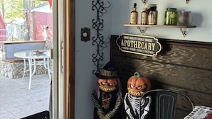 Une pièce avec une table en bois, une chaise noire, un mur décoratif orné d’une enseigne indiquant « PHARMACIE », et des décorations d’Halloween, notamment deux citrouilles sculptées.