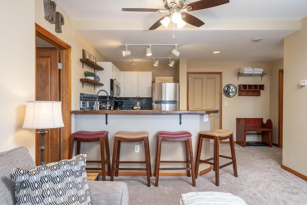 A compact kitchen with a central island, bar stools, a ceiling fan, and a dining area with a wooden bench.