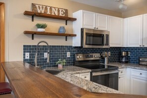 A kitchen with white cabinets, a stainless steel microwave, a stove, and a sink. There are wooden shelves above the sink with decorative items.