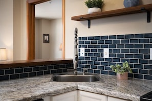 A kitchen with a granite countertop, a stainless steel sink, a blue-tiled backsplash, and a wooden shelf above.