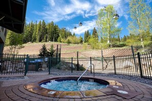 A hot tub surrounded by a brick patio, a fenced area, and a backdrop of trees and a ski lift.