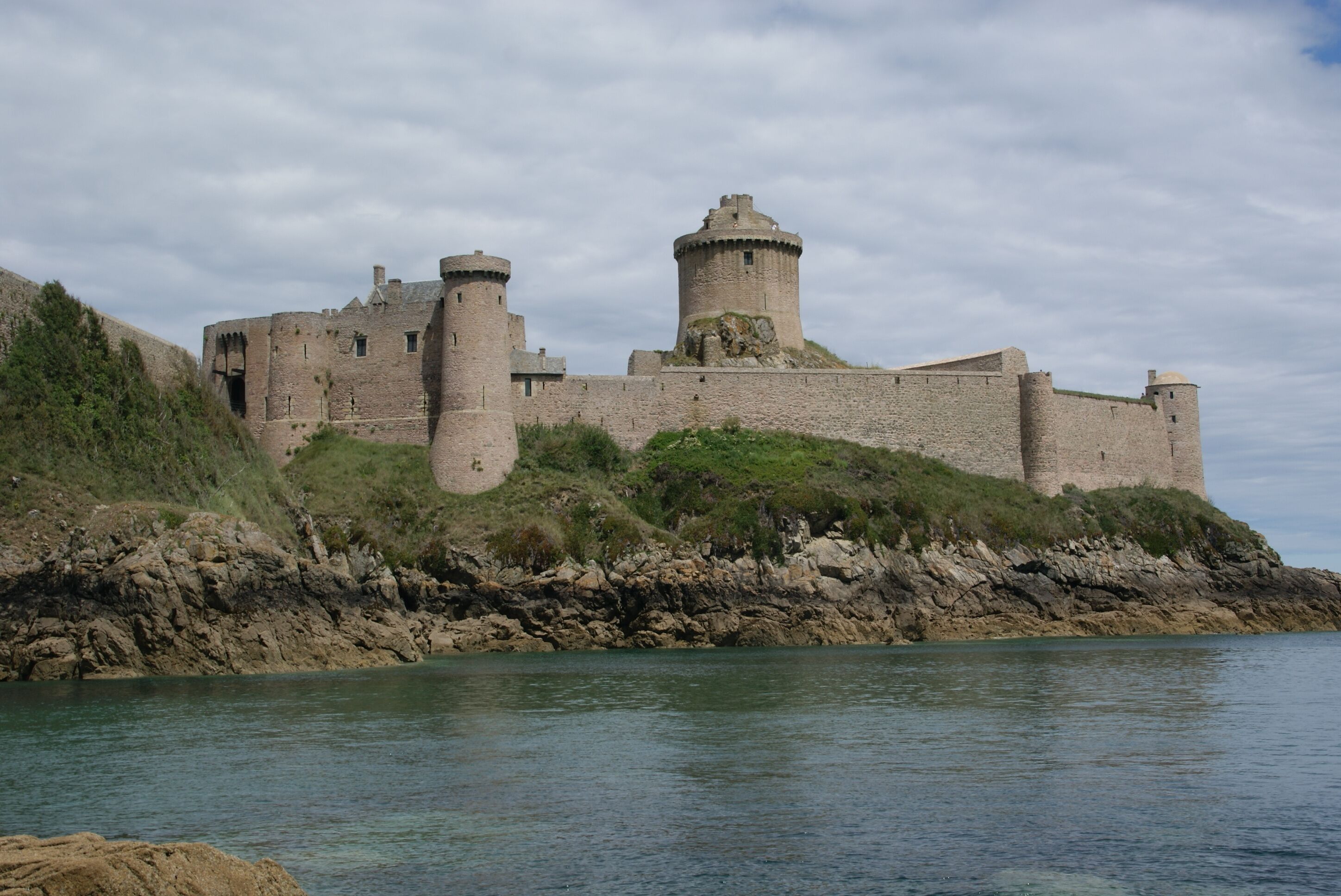 Eine historische Steinburg mit mehreren Türmen, gelegen an einer felsigen Küste, im Vordergrund klares Wasser.