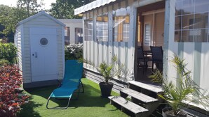 A small, single-story building with a covered porch, a blue lawn chair, and a wooden bench. - Top-of-the-range mobile home (Dinard)