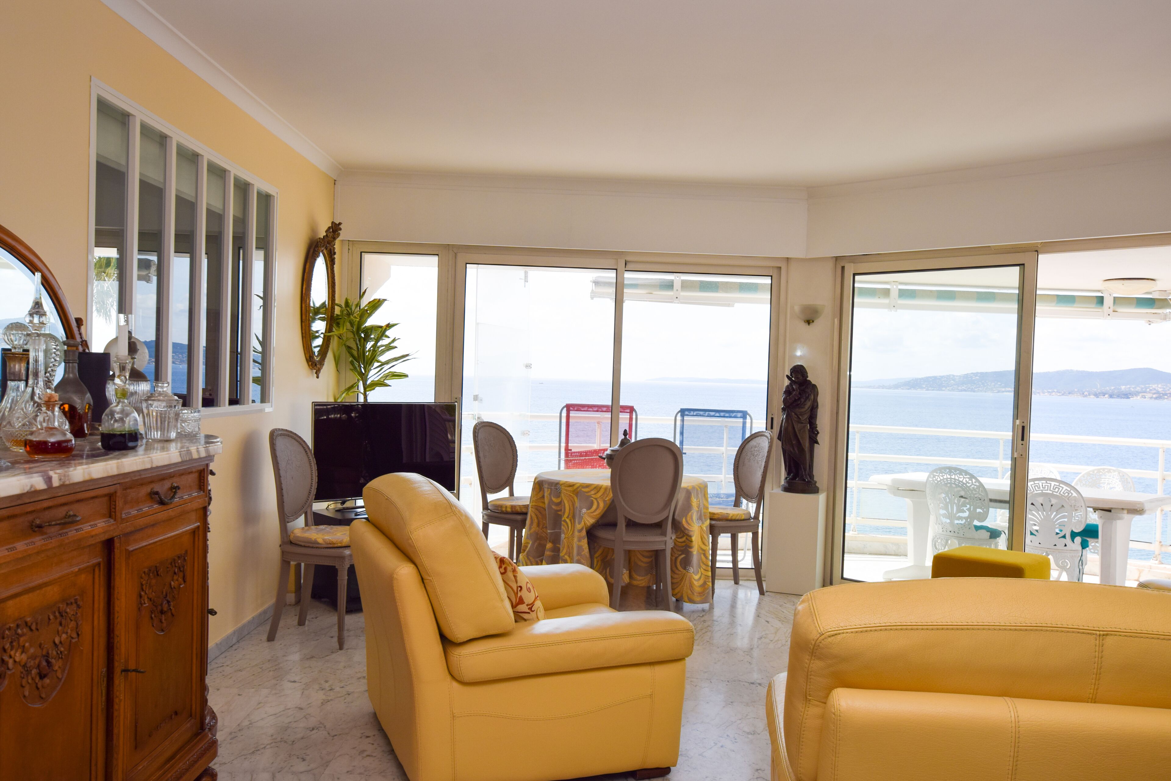Een kamer met een geel sofa, een televisie, een eettafel met stoelen en uitzicht op zee door grote ramen.