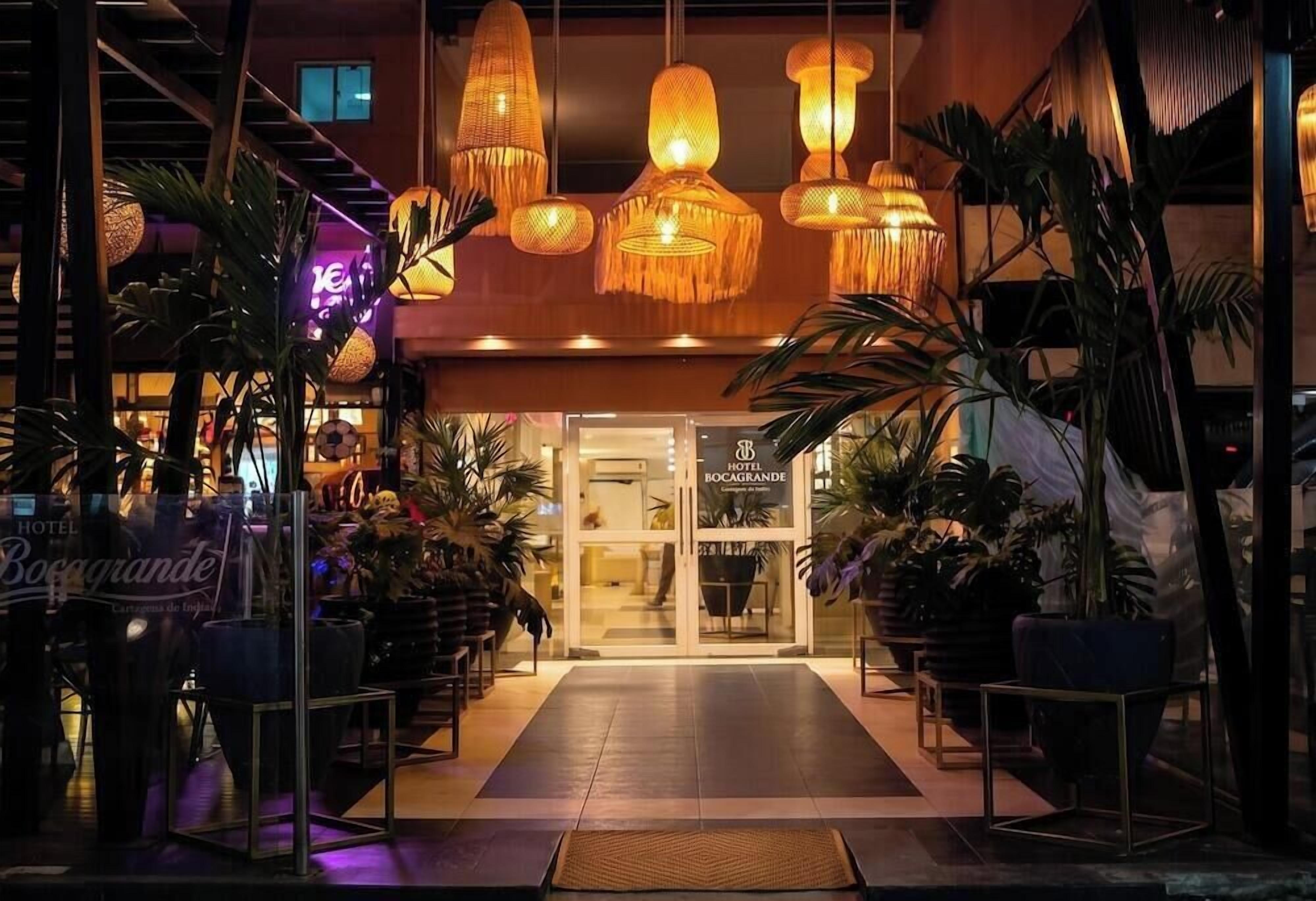 Entrada de hotel con diseño moderno, que incluye puerta de vidrio, plantas en macetas y lámparas colgantes.