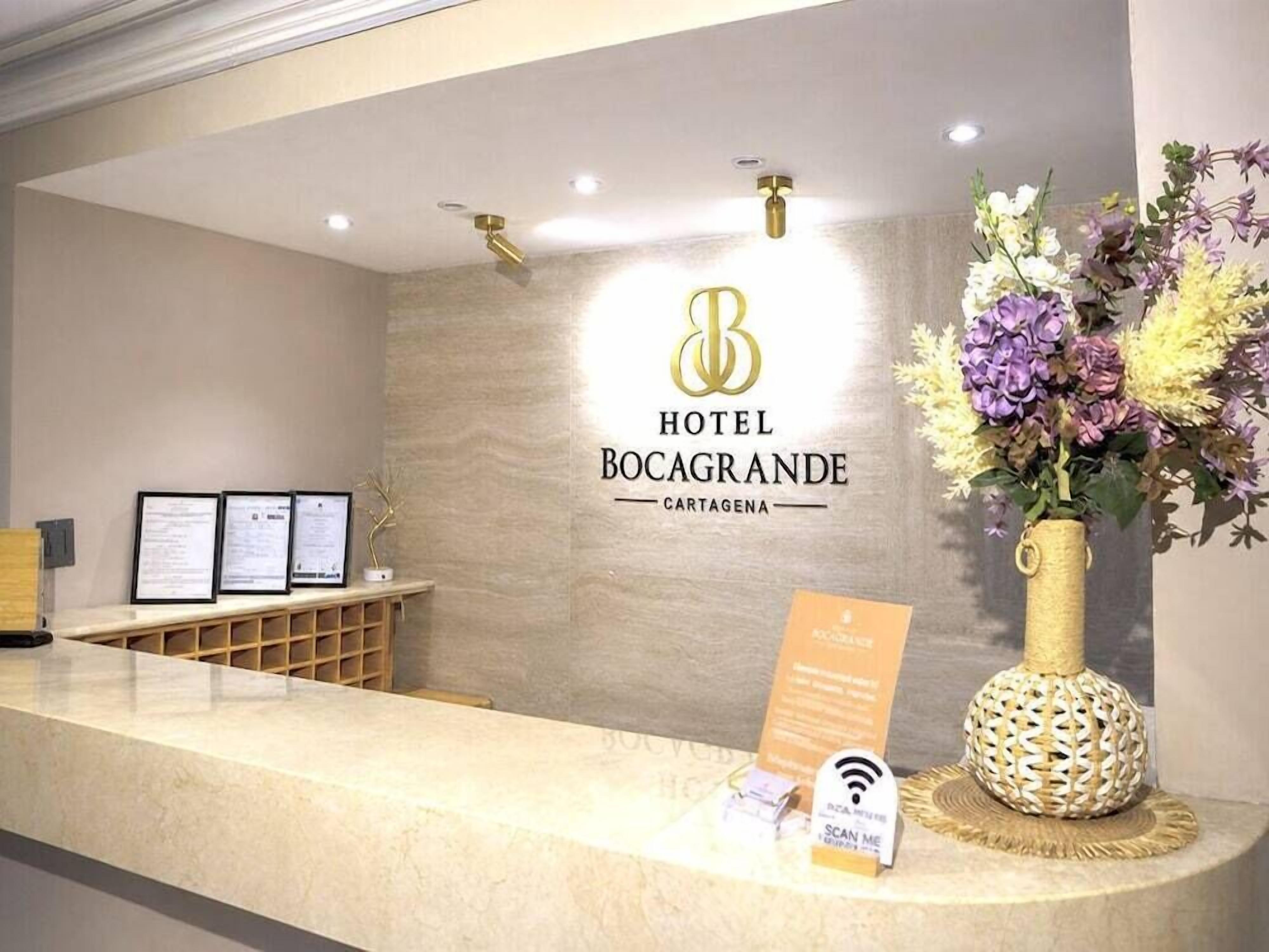 Un mostrador de recepción de hotel con un logotipo, documentos enmarcados, un jarrón de flores y un cartel de Wi-Fi.