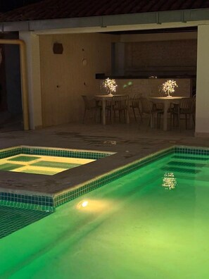 A swimming pool with a tiled edge, a dining area with chairs, and a kitchen area with a countertop.