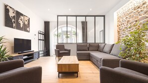 Uma sala de estar moderna com um sofá seccional, uma mesa de centro de madeira e uma TV sobre um suporte. Há uma janela grande com cortinas, uma parede de pedra e uma planta.