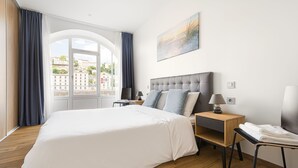 Quarto de hotel moderno com uma cama grande, uma mesa de cabeceira de madeira, uma escrivaninha com abajur e vista para a cidade através de uma janela ampla.