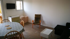 A living room with a round table, chairs, a sofa, a TV mounted on the wall, and a window providing natural light. - Self-contained apartment, patio with terrace and bicycle paths (La Teste-de-Buch)