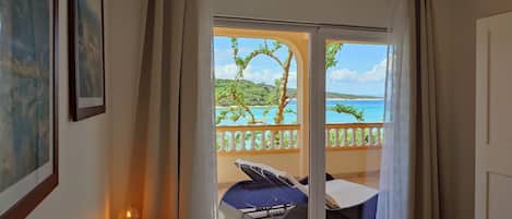 A hotel room with a bed, a small table with a lamp, and a view of a beach and ocean through a sliding door.
