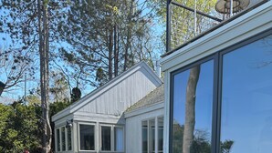 Une terrasse extérieure couverte, aménagée avec des meubles en osier et une table en verre, entourée d’une terrasse en bois et d’un bâtiment blanc doté de grandes fenêtres.