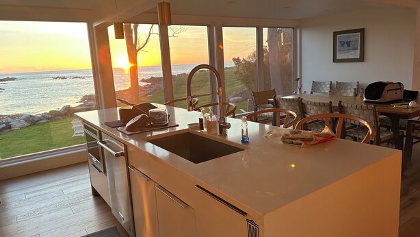 Une cuisine moderne dotée d’un grand îlot central, d’un évier et offrant une vue sur l’océan au coucher du soleil.