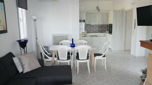 A modern living room with a black sofa, white dining table, and chairs, a kitchen in the background, and a TV mounted on the wall. - George place two-storey holiday home offers panoramic sea and mountain views. (Λυγαριά)