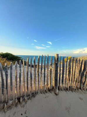 A wooden fence with barbed wire, leading to a sandy beach with the ocean in the background.