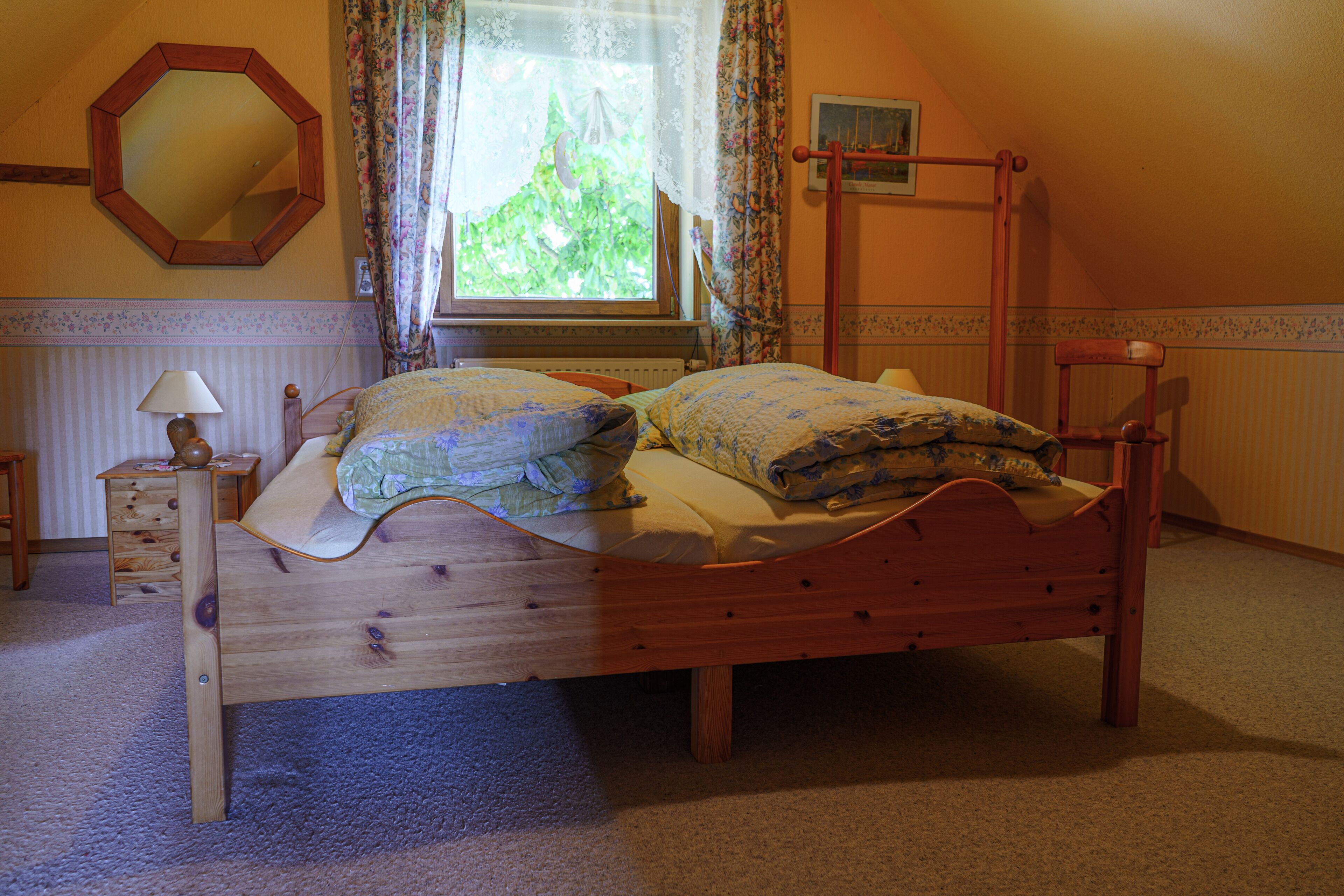 Een slaapkamer met een houten bed, nachtkastje, lamp, raam met gordijnen en een spiegel aan de muur.